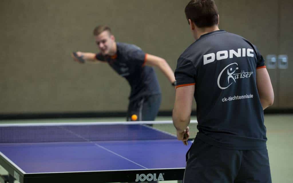 Aktiver Tischtennissport bei CK-Tischtennis: Trainer beobachtet Spieltechnik, während Spieler einen Topspin ausführt, auf einem hochwertigen Joola Tisch. Perfektes Training für Hobby und Leistungssport.