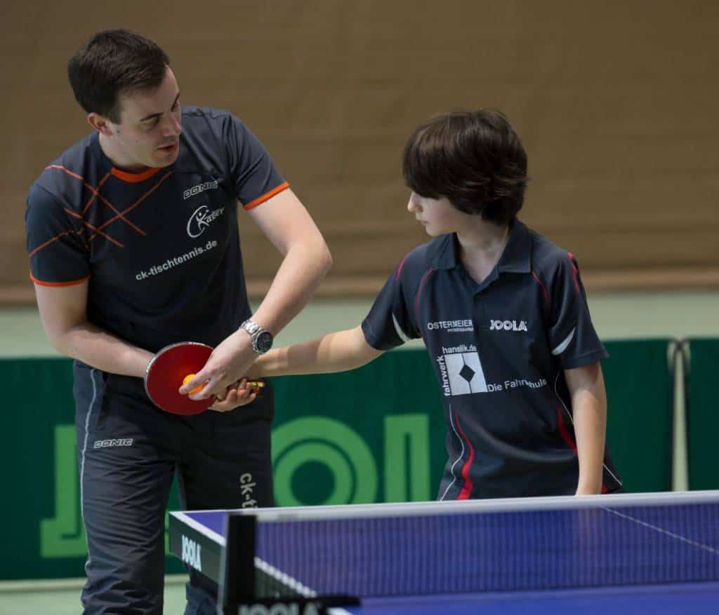 Lernender Tischtennisspieler erhält Anweisung von Trainer beim Training, vom Tischtennisverein CK-Tischtennis, in der Halle, professionelle Schlägerhaltung, Training, Unterstützung, Sport, Kinder und Jugendliche.