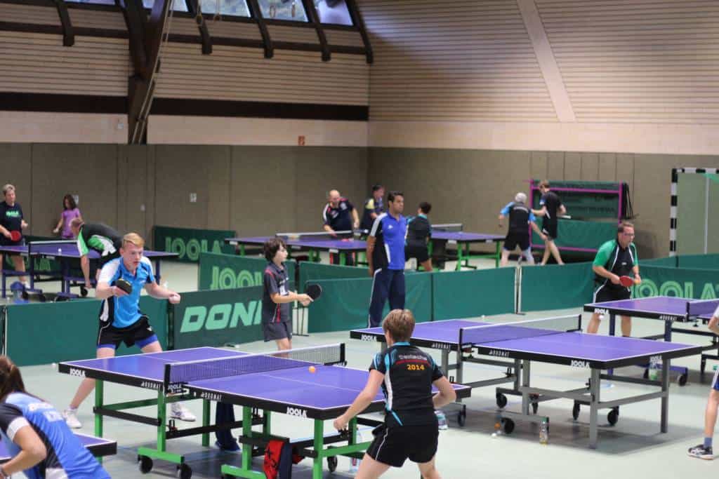 Modernes Tischtennistraining in einer Indoor-Halle mit mehreren Tischen und Spielern unterschiedlichen Alters, ideal für Tischtennis-Training und -Wettkämpfe im Verein.